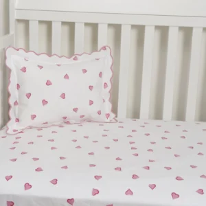 Crib with white bedding featuring pink heart patterns and a matching scalloped pillow.