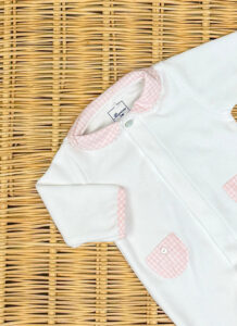 White baby onesie with pink gingham collar and pockets, placed on a woven surface.
