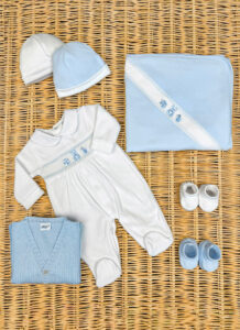 A collection of baby clothing: two hats, a onesie, a blanket, a sweater, and two pairs of shoes, all in blue and white.