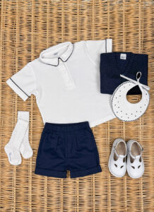 A white polo shirt, navy shorts, a navy sweater, socks, a bib, and white shoes arranged on a woven surface.
