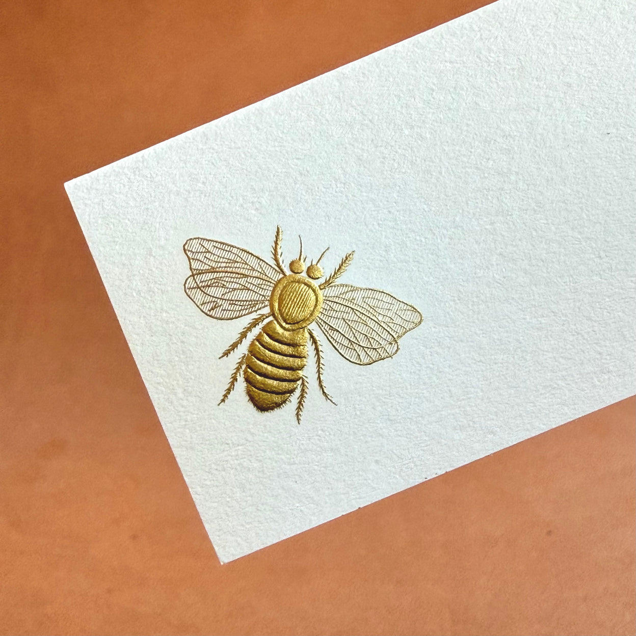 A detailed illustration of a golden bee on textured white card, set against a warm brown background.