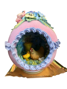 A decorative sugar egg features a hen and chick inside, surrounded by pastel flowers and grass.